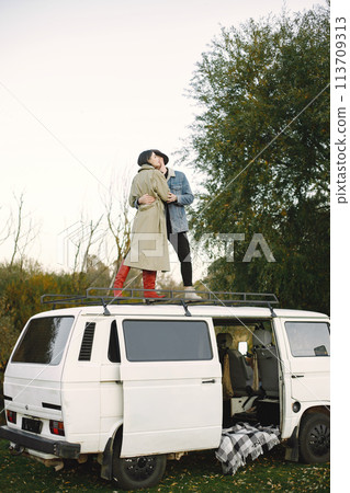 Man and woman in fashion clothes standing on top of the bus. Man wearing jacket and a black hat and woman coat and red boots. Photo of romantic couple on the top of retro bus. 113709313