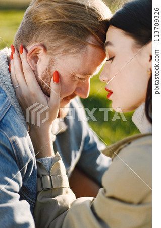 Man and woman in fashion clothes sitting on a nature on a picnic rug. Man wearing jacket and woman coat. Woman looking at man. Man and woman in fashion clothes sitting on a nature on a picnic rug. Man wearing jacket and woman coat. Woman looking at man. 113709326