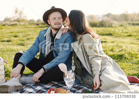 Man and woman in fashion clothes sitting on a nature on a picnic rug. Man wearing jacket and a black hat and woman coat. Woman hugging a man, Man and woman in fashion clothes sitting on a nature on a picnic rug. Man wearing jacket and a black hat and woman coat. Woman hugging a man, 113709330