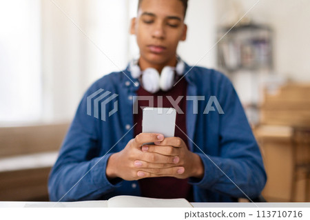 Focused black teen guy using phone with headphones around neck 113710716