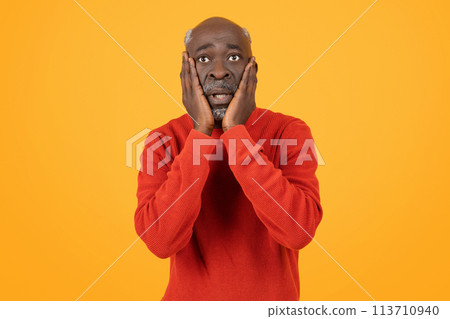 Shocked senior black man with hands on cheeks and wide eyes, wearing a red sweater 113710940