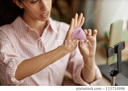Close up of young woman holding pink menstrual cup while recording educational videos via smartphone Close up of young woman holding pink menstrual cup while recording educational videos via smartphone 113710996