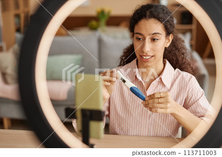 Portrait of smiling young woman holding pregnancy test stick while recording female health educational videos via smartphone with ring light copy space 113711003