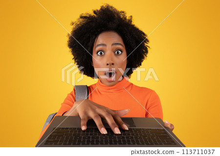 Surprised African American woman with curly hair wearing an orange turtleneck is staring at her laptop. Surprised African American woman with curly hair wearing an orange turtleneck is staring at her laptop. 113711037