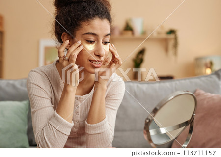 Portrait of smiling young woman applying moisturizing patches and looking in mirror at home 113711157
