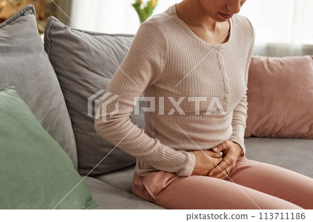 Close up of unrecognizable young woman holding onto stomach suffering from cramps sitting on couch at home copy space Close up of unrecognizable young woman holding onto stomach suffering from cramps sitting on couch at home copy space 113711186