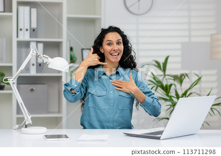 Joyful businesswoman with curly hair in a blue shirt sits at her office desk, gesturing a 'call me' sign and smiling energetically. Joyful businesswoman with curly hair in a blue shirt sits at her office desk, gesturing a 'call me' sign and smiling energetically. 113711298