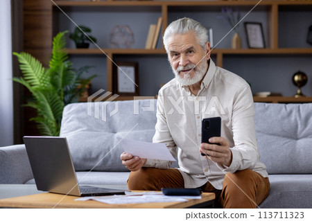 Elderly bearded man sitting on a couch, holding documents and smartphone while working with a laptop. Comfortable home office setup in a cozy living room. Elderly bearded man sitting on a couch, holding documents and smartphone while working with a laptop. Comfortable home office setup in a cozy living room. 113711323