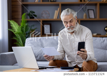 Focused elderly man with white hair and beard, reviewing papers and checking information on his smartphone while sitting on a couch. A comfortable home setting with laptop and indoor plants. Focused elderly man with white hair and beard, reviewing papers and checking information on his smartphone while sitting on a couch. A comfortable home setting with laptop and indoor plants. 113711324