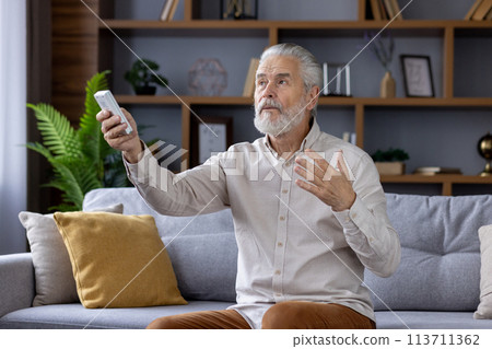Elegant senior man sitting on a gray couch, comfortably using a remote control to operate the air conditioning in his stylish, contemporary living room. 113711362