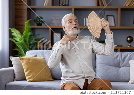Elderly caucasian man sitting on a gray sofa, using a handheld fan to cool off during a warm day indoors, looking uncomfortable and sweaty in a well-decorated living room. Elderly caucasian man sitting on a gray sofa, using a handheld fan to cool off during a warm day indoors, looking uncomfortable and sweaty in a well-decorated living room. 113711367