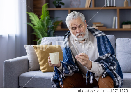 Elderly Caucasian man wrapped in a blanket, sitting on a couch and checking his fever with a thermometer. He holds a mug, looking worried and unwell in his living room. 113711373