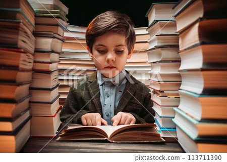 Cute little schoolboy reading book in library between stacks of books literature 113711390