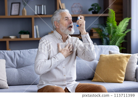 Elderly Caucasian man experiencing an asthma attack, using a blue inhaler while sitting on a couch in a modern living room. Elderly Caucasian man experiencing an asthma attack, using a blue inhaler while sitting on a couch in a modern living room. 113711399