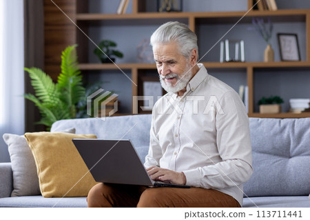 A cheerful senior man with gray hair sits comfortably on a sofa, deeply engaged in using his laptop within a modern living room decorated with indoor plants. A cheerful senior man with gray hair sits comfortably on a sofa, deeply engaged in using his laptop within a modern living room decorated with indoor plants. 113711441