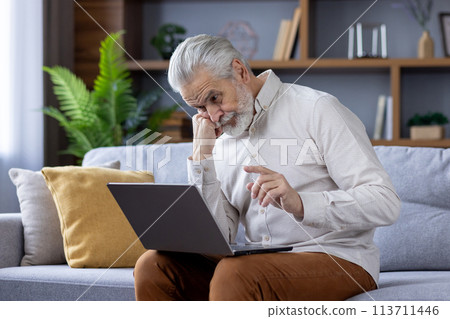 Focused elderly man with white hair and beard deeply engrossed in using a laptop while sitting on a sofa in a well-lit living room, depicting modern technology use by seniors. Focused elderly man with white hair and beard deeply engrossed in using a laptop while sitting on a sofa in a well-lit living room, depicting modern technology use by seniors. 113711446