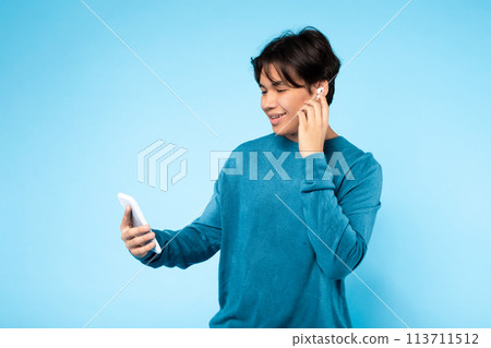 Young guy using earbuds with smartphone, blue background Young guy using earbuds with smartphone, blue background 113711512