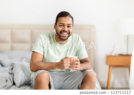 Cheerful african american man with coffee sitting on bed 113711517