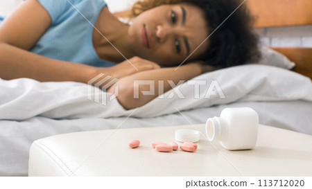 Sick woman looking at pills, lying on bed 113712020