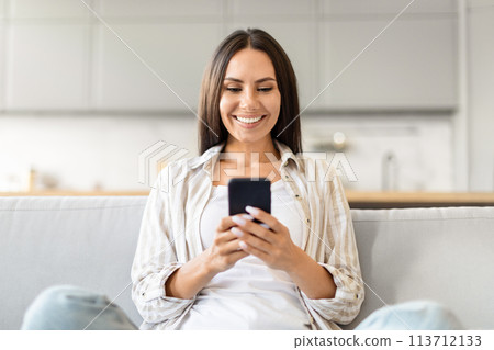 Confident woman using smartphone and smiling at home 113712133