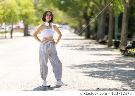 A woman is standing on a sidewalk wearing headphones and a white tank top A woman is standing on a sidewalk wearing headphones and a white tank top 113712173