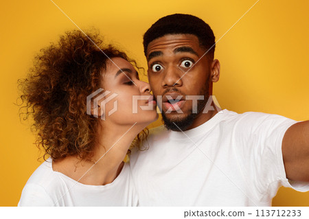 Shocked african guy taking selfie, woman kissing his cheek 113712233