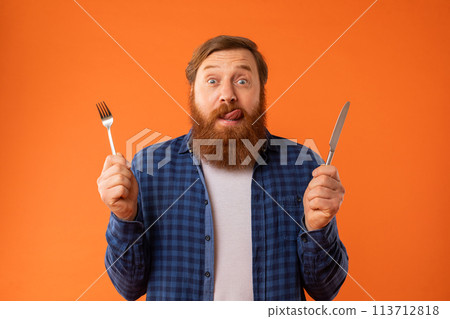 Hungry redhaired bearded guy holding knife and fork, orange background Hungry redhaired bearded guy holding knife and fork, orange background 113712818