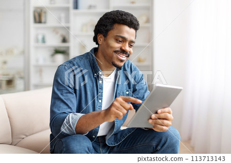 Smiling Black Man Using Digital Tablet On Couch in Bright Living Room Smiling Black Man Using Digital Tablet On Couch in Bright Living Room 113713143