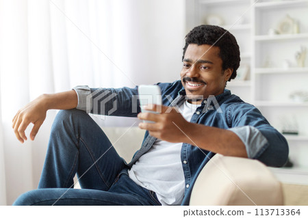 Smiling Black Man Enjoying Time Indoors With His Smartphone Smiling Black Man Enjoying Time Indoors With His Smartphone 113713364