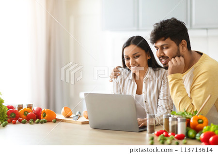 Happy loving young indian spouses using laptop while cooking Happy loving young indian spouses using laptop while cooking 113713614