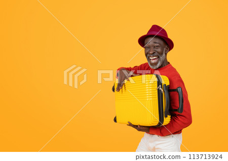Cheerful senior black man wearing a red hat and sweater, holding a bright yellow suitcase 113713924