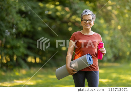 Senior woman holding water bottle and yoga mat, ready for training outdoors Senior woman holding water bottle and yoga mat, ready for training outdoors 113713971