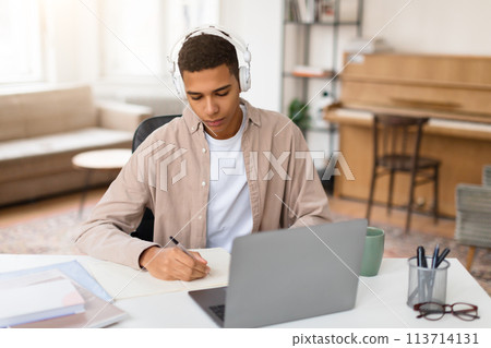 Man with headphones writing notes at desk Man with headphones writing notes at desk 113714131