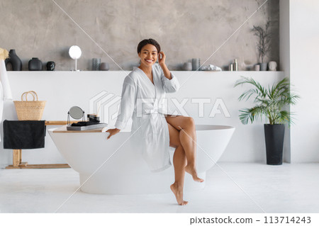 Smiling black woman in bathrobe sitting on edge of luxurious freestanding bathtub 113714243
