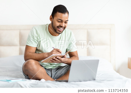 Man taking notes with laptop on bed 113714350