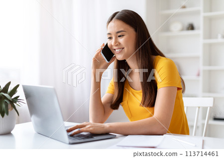 Happy asian woman talking on cellphone while working on laptop at home 113714461