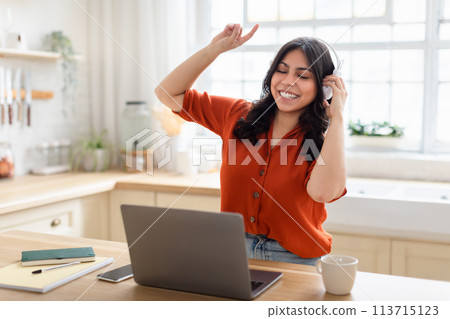 Joyful woman dancing with headphones in front of laptop 113715123