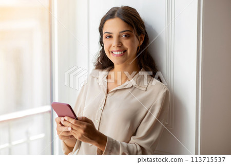 Happy young woman holding smartphone and smiling while standing by window 113715537