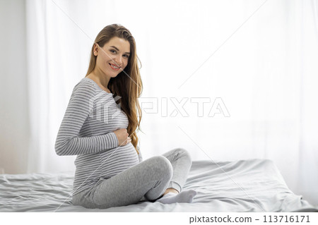 Happy pregnant woman sitting on bed at home Happy pregnant woman sitting on bed at home 113716171