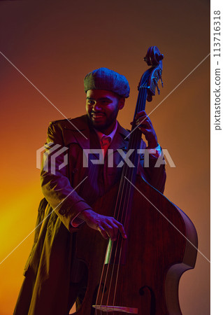 African-American man, musician in vintage coat and cap creates rhythm on bass in neon light against gradient background. 113716318