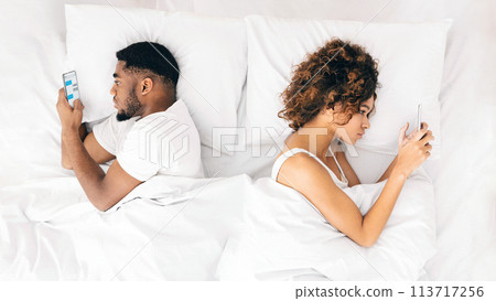Millennial couple lying on bed back to back with smartphones Millennial couple lying on bed back to back with smartphones 113717256