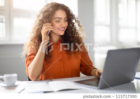 Woman conversing on phone with laptop at office Woman conversing on phone with laptop at office 113717300