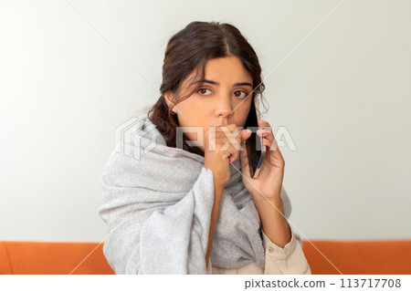 Coughing young woman wrapped in blanket talking on cellphone at home Coughing young woman wrapped in blanket talking on cellphone at home 113717708