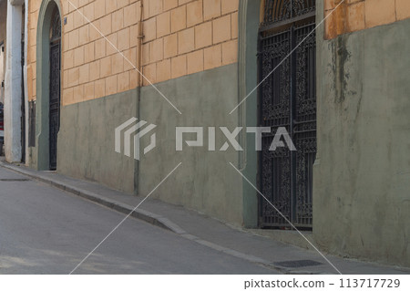 A city street with a perspective of a sidewalk stretching into the distance, a roadway, a building wall with a large massive metal gate with forged elements and patterns A city street with a perspective of a sidewalk stretching into the distance, a roadway, a building wall with a large massive metal gate with forged elements and patterns 113717729