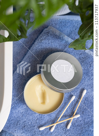 Opened lip balm jar on blue towels near basin and green monstera in bath, lid mockup 113717937