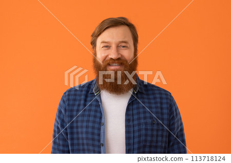 Redhaired bearded man stands smiling to camera against orange background 113718124