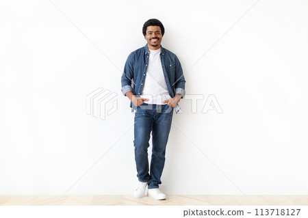 Cheerful black man standing confidently with hands in pockets against white wall Cheerful black man standing confidently with hands in pockets against white wall 113718127