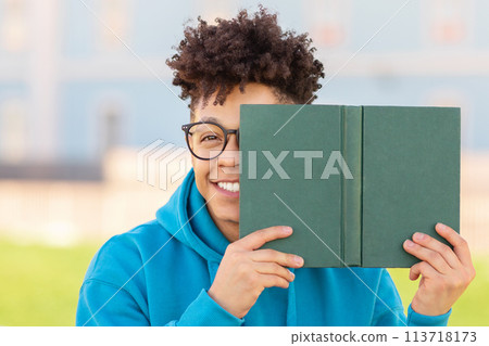 Playful student hiding behind book outdoors 113718173