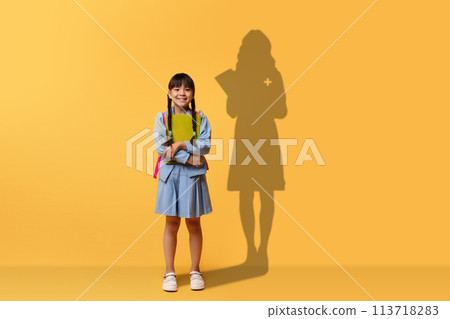 A cheerful young girl clasps a book and smiles, wearing a blue dress and backpack A cheerful young girl clasps a book and smiles, wearing a blue dress and backpack 113718283