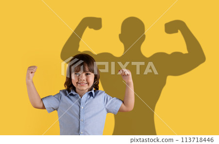 A smiling child in a blue shirt shows off muscles with a superhero shadow in the background A smiling child in a blue shirt shows off muscles with a superhero shadow in the background 113718446